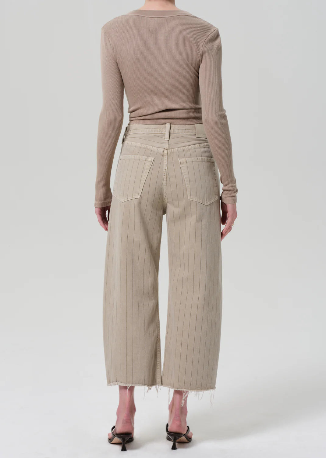 A person wears a fitted, long-sleeve beige top with Citizens Of Humanity/AGOLDE Ayla Raw Hem Crop high-waisted, wide-leg pinstriped pants in rigid cotton, relaxed rise, standing with their back to the camera against a plain light background.