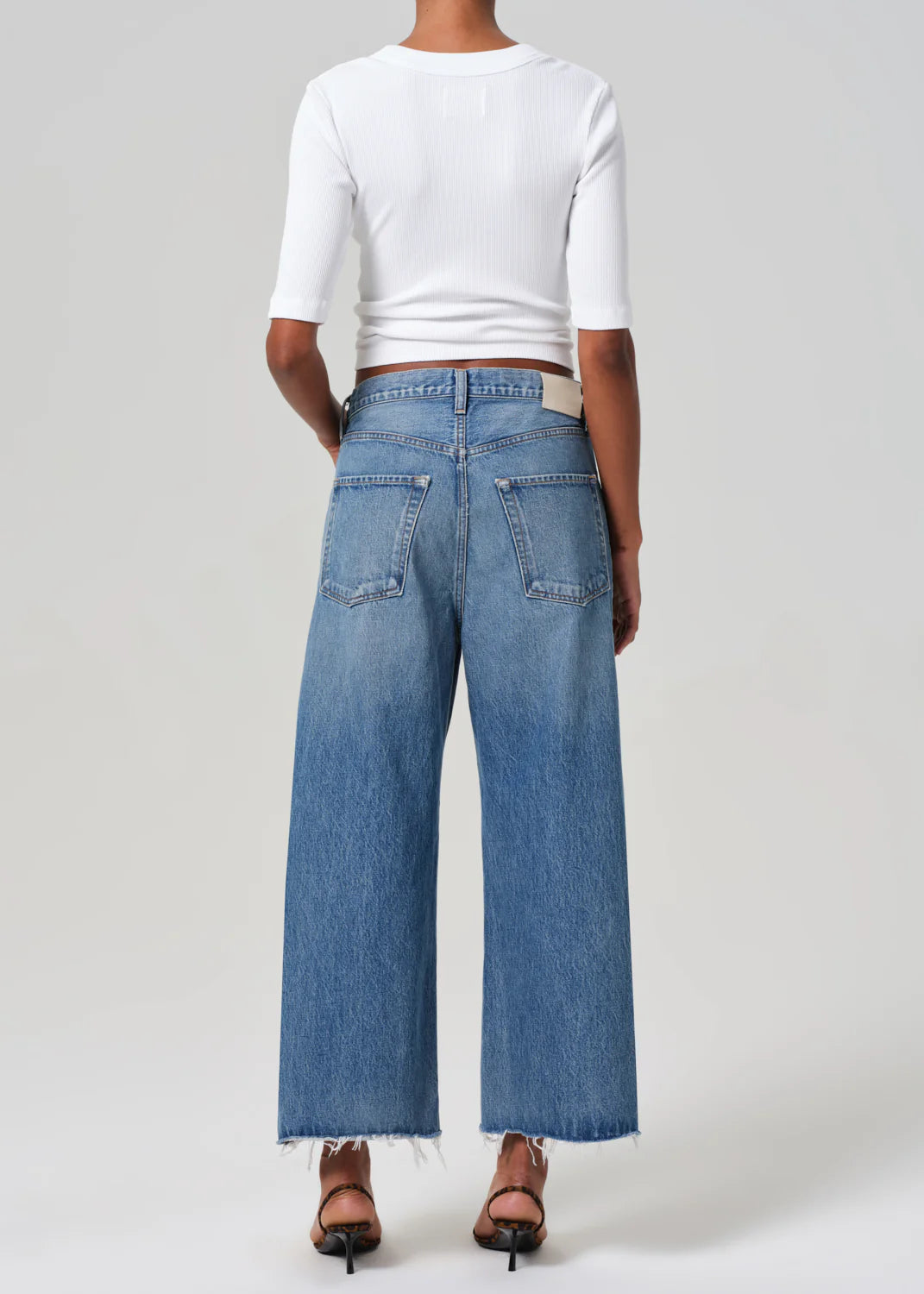 From the back, a person wears Citizens Of Humanity's Ayla Raw Hem Crop and a white ribbed three-quarter sleeve top, creating a vintage silhouette with wide-leg jeans. They also sport open-toed black heeled sandals on a plain backdrop.