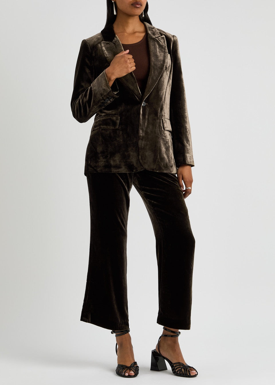 A person wearing a Slim Velvet Blazer by FRAME featuring peaked lapels and coordinating wide-leg pants pairs the outfit with a brown top and open-toe high-heeled sandals. They stand against a plain white background, exuding elegance and style.