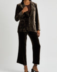 A person wearing a Slim Velvet Blazer by FRAME featuring peaked lapels and coordinating wide-leg pants pairs the outfit with a brown top and open-toe high-heeled sandals. They stand against a plain white background, exuding elegance and style.