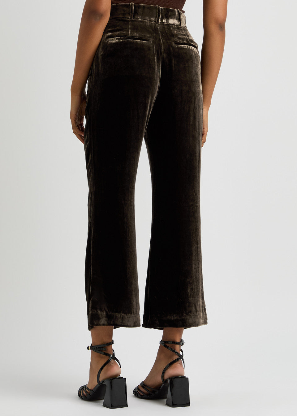 A rear view of a person wearing the Crop Flare Velvet Trouser by FRAME in dark brown velvet, paired with black strappy high-heeled sandals. These trousers boast a high-waisted design and feature wide-leg cropped pants, set against a plain white background.