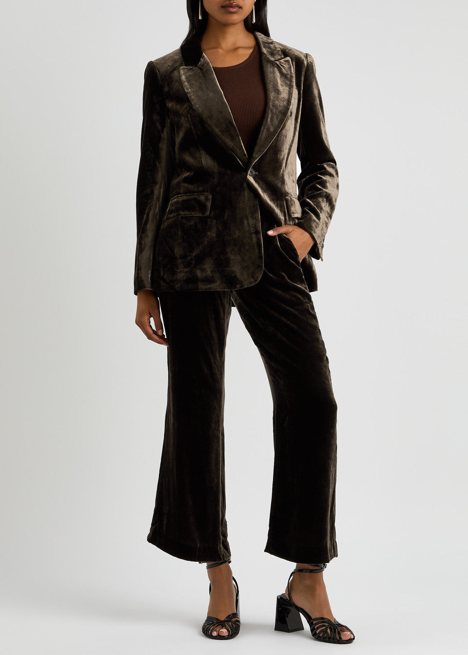 A person dressed in FRAME's Crop Flare Velvet Trouser and a matching blazer stands against a plain background. The outfit is enhanced with a brown top and black open-toe heels.