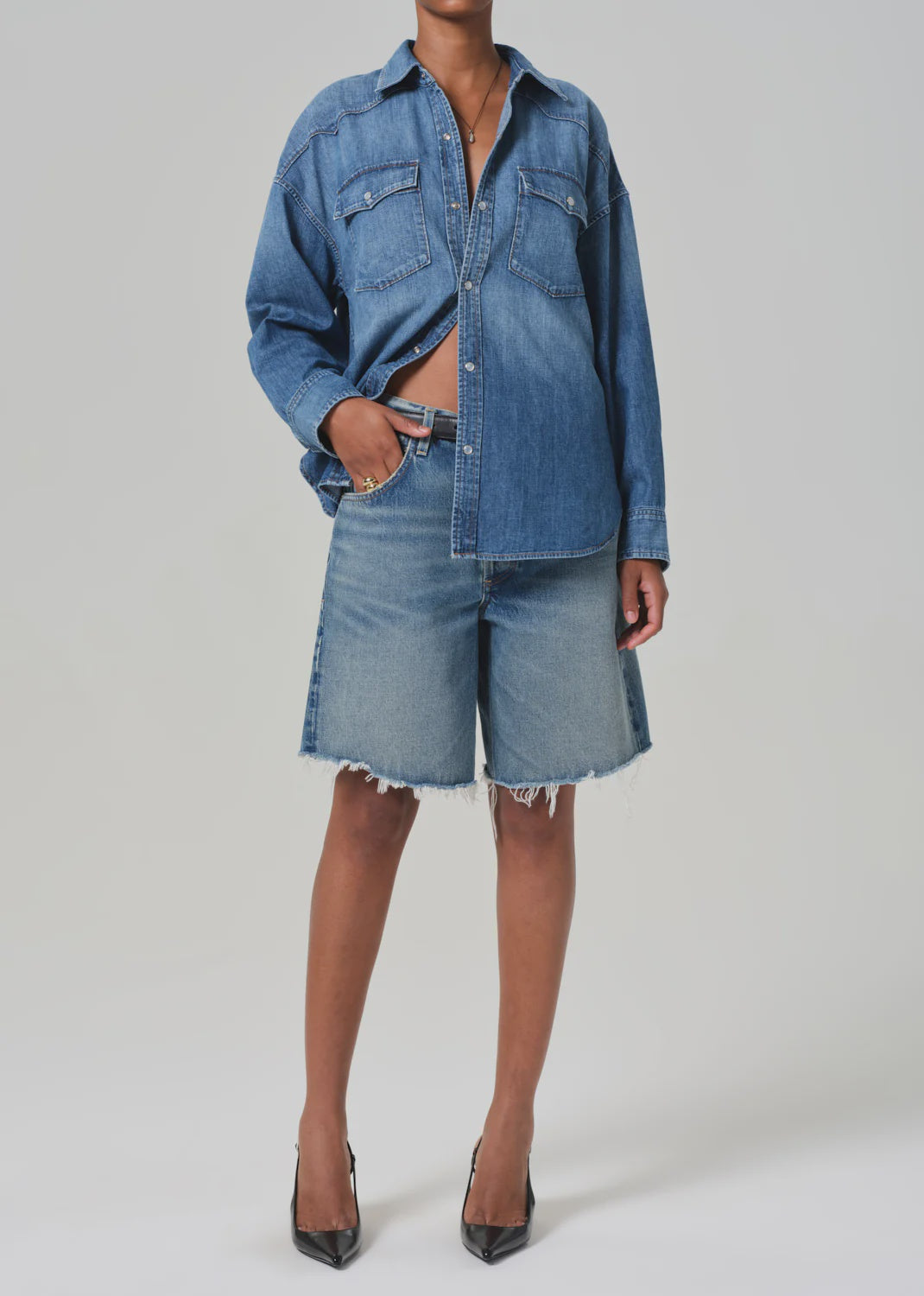 Against a gray backdrop, someone models Citizens Of Humanity/AGOLDE's Ayla Short and a loose denim shirting. With one hand in their pocket and black pointed heels, the look showcases casual elegance and relaxed style using regenerative cotton.