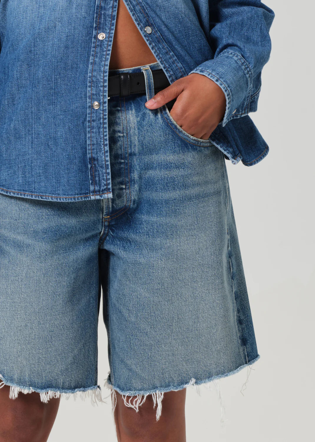 Against a light background, someone wearing a medium indigo denim shirt made from regenerative cotton and AGOLDE's Ayla Short with a black belt stands. The shirt is partially unbuttoned, while their hand rests casually in the versatile inseam pocket of the shorts.