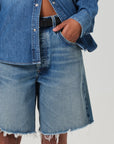 Against a light background, someone wearing a medium indigo denim shirt made from regenerative cotton and AGOLDE's Ayla Short with a black belt stands. The shirt is partially unbuttoned, while their hand rests casually in the versatile inseam pocket of the shorts.