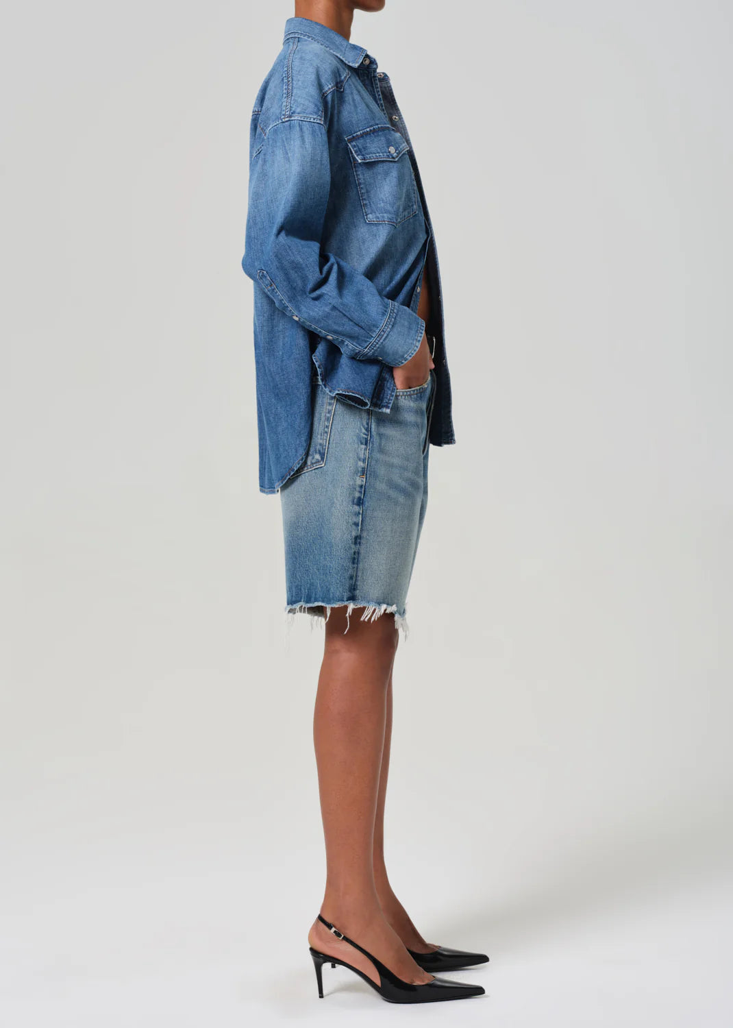 A person stands in profile wearing Citizens Of Humanity/AGOLDE's Ayla Short in medium indigo denim, crafted from regenerative cotton, paired with a frayed denim skirt. They complete the look with black sling-back heels against a plain gray background.