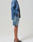 A person stands in profile wearing Citizens Of Humanity/AGOLDE's Ayla Short in medium indigo denim, crafted from regenerative cotton, paired with a frayed denim skirt. They complete the look with black sling-back heels against a plain gray background.