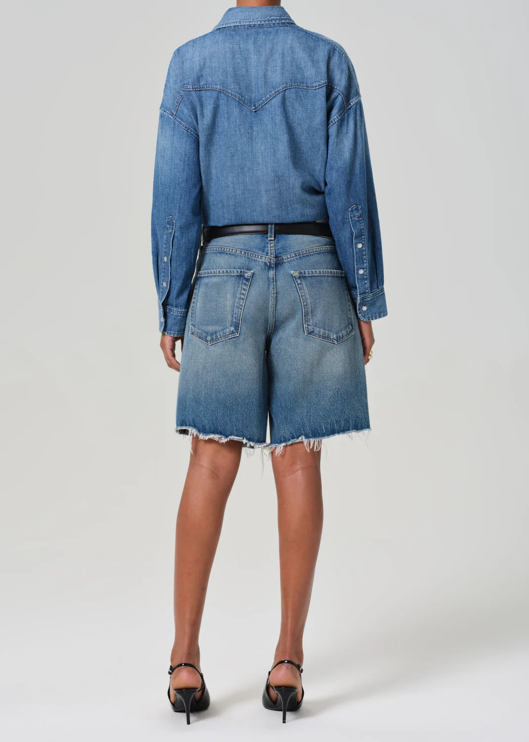 A rear view showcases a person wearing an Ayla Baggy long-sleeve denim shirt and Ayla Shorts by Citizens Of Humanity/AGOLDE. Crafted from regenerative cotton, these high-waisted shorts with frayed hems are styled with black heels against a plain backdrop, emphasizing their versatile inseam.