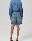 A rear view showcases a person wearing an Ayla Baggy long-sleeve denim shirt and Ayla Shorts by Citizens Of Humanity/AGOLDE. Crafted from regenerative cotton, these high-waisted shorts with frayed hems are styled with black heels against a plain backdrop, emphasizing their versatile inseam.