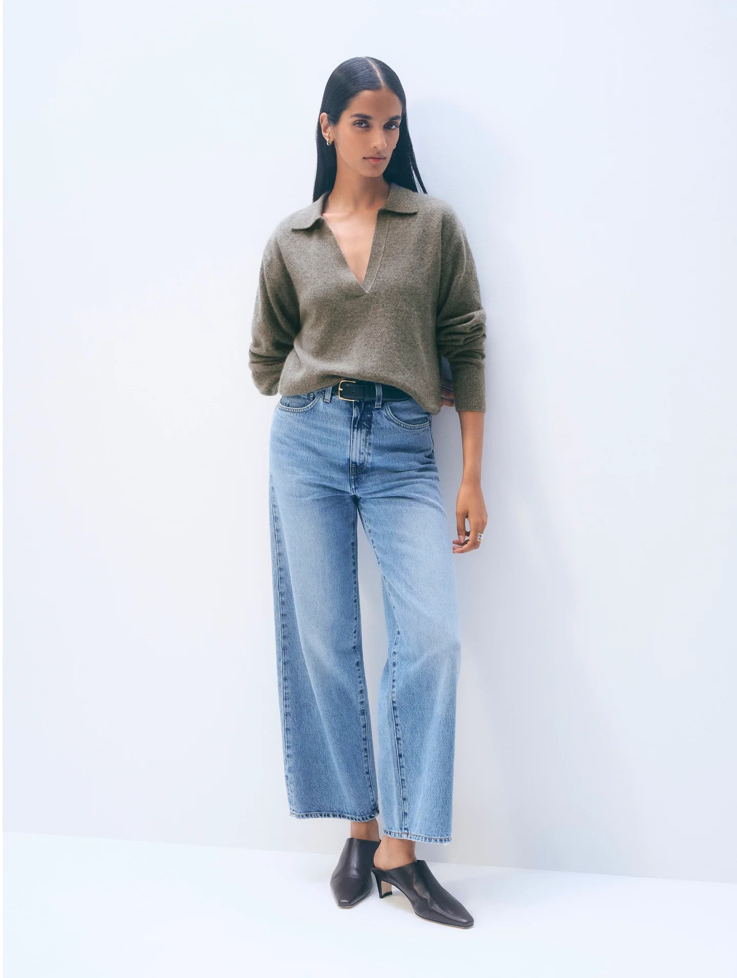 A woman with long straight dark hair and a neutral expression stands against a plain light background, wearing the White + Warren Cashmere Polo Top in brown, high-waisted wide-leg blue jeans, and pointed dark brown shoes.