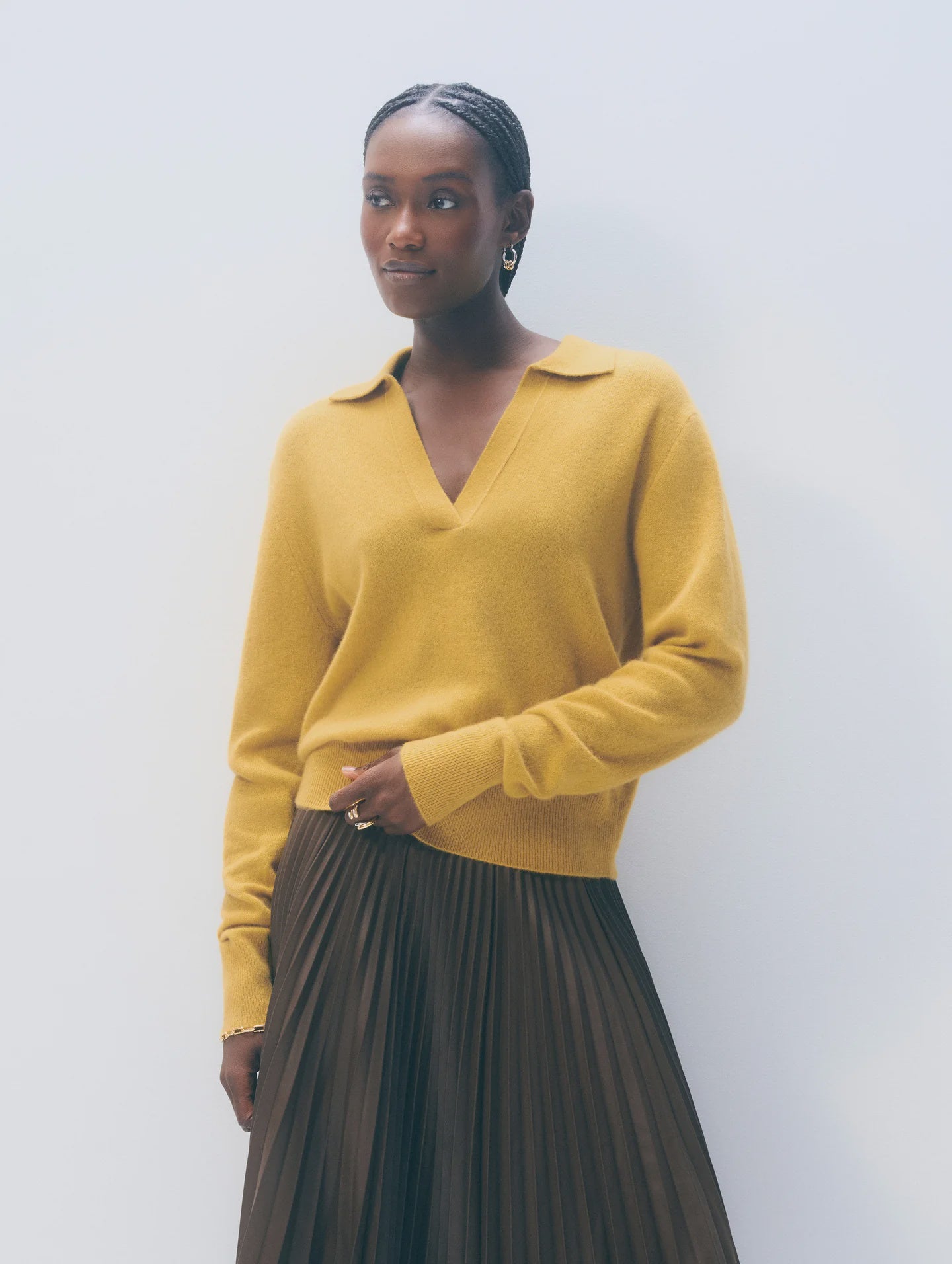A woman with short braided hair and small hoop earrings stands against a light background, wearing the White + Warren Cashmere Polo Top in mustard yellow with a collar and a dark pleated skirt.