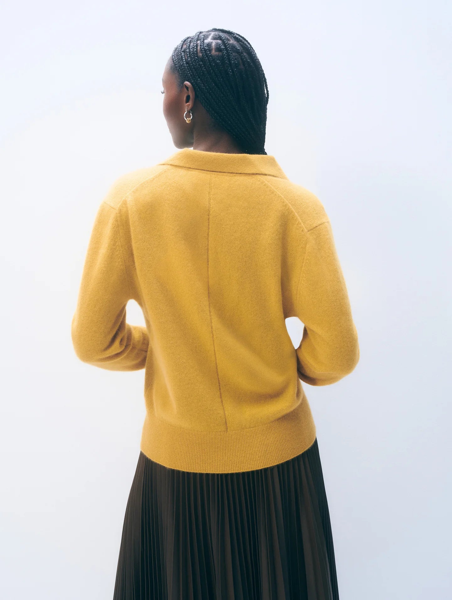 A person with braided hair, wearing the White + Warren Cashmere Polo Top in mustard yellow and a dark pleated skirt, stands facing away from the camera against a plain light background.