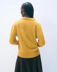 A person with braided hair, wearing the White + Warren Cashmere Polo Top in mustard yellow and a dark pleated skirt, stands facing away from the camera against a plain light background.