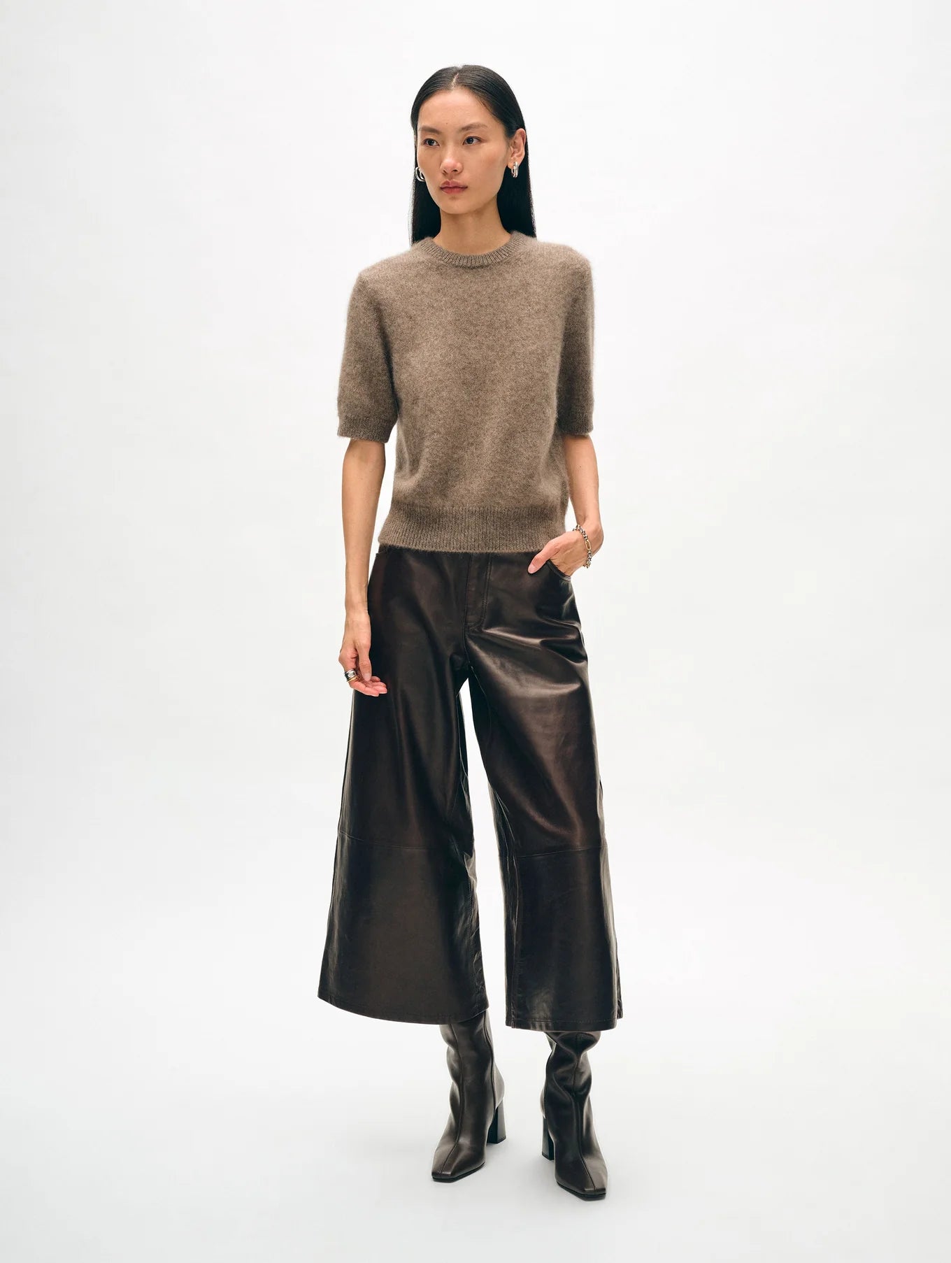 A person stands against a plain background, exuding luxurious comfort in a Brushed Cashmere Tee from White + Warren and dark brown wide-leg leather pants. Paired with brown high-heeled boots, this versatile ensemble boasts a stylish and modern appearance.