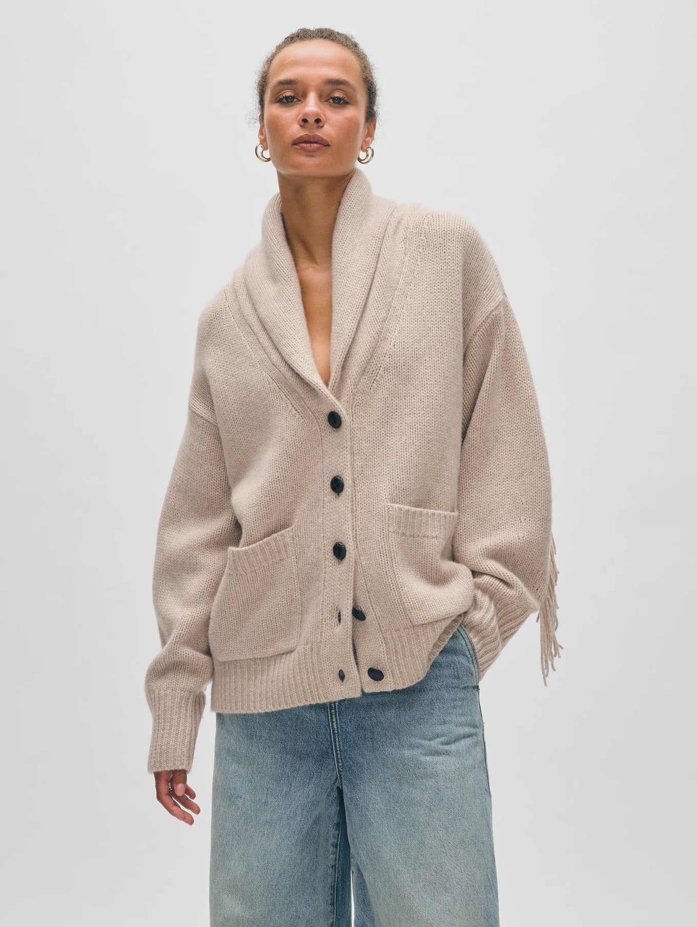 A model in light blue jeans and the White + Warren Cashmere Blend Luxe Fringe Coatigan, a beige merino wool-cashmere button-up cardigan with large front pockets, thick collar, and fringe sleeves, poses against a plain white background.