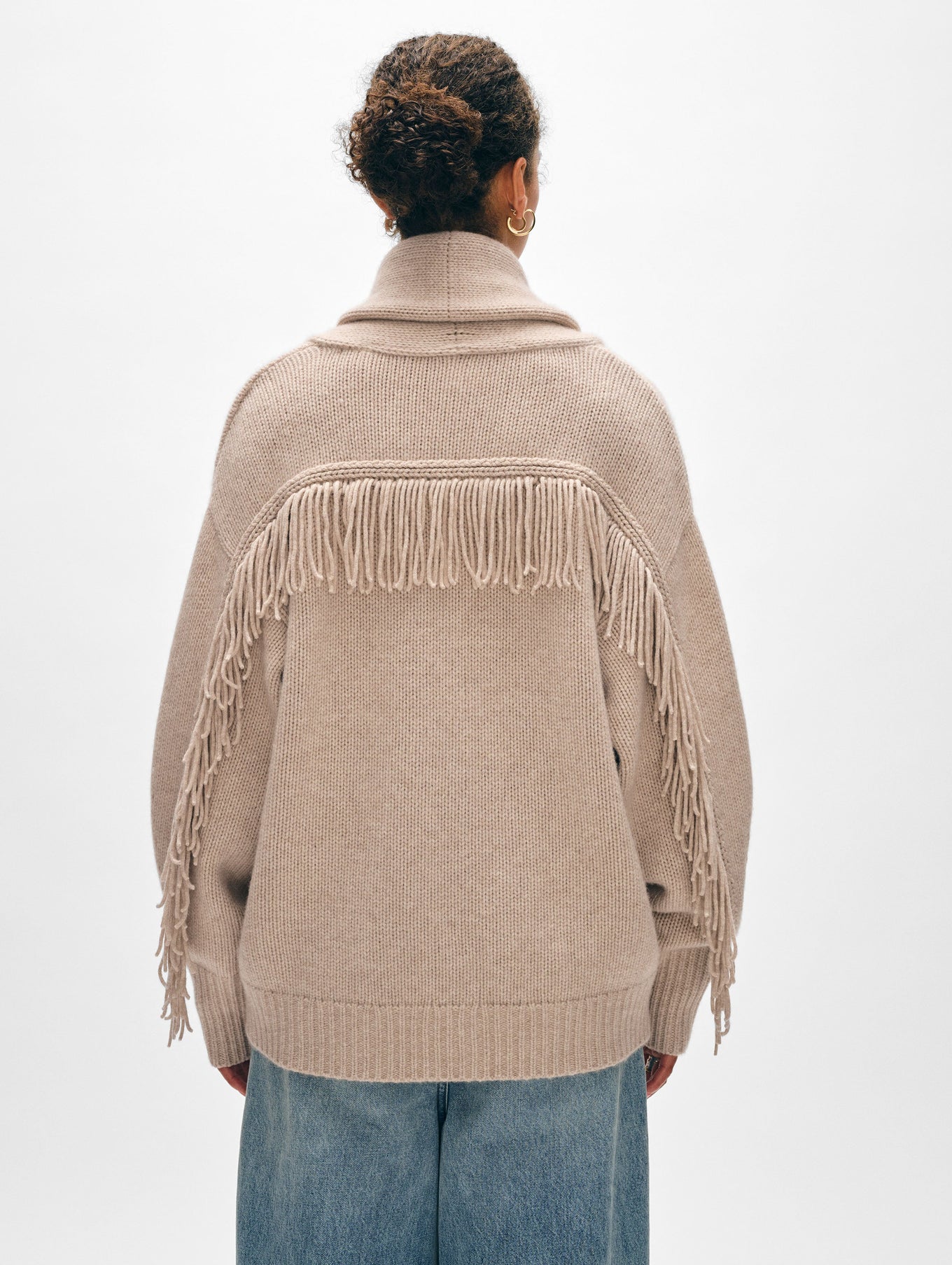 A person poses facing away in a White + Warren Cashmere Blend Luxe Fringe Coatigan and blue denim jeans. The beige coatigan, crafted from a luxurious merino wool-cashmere blend, features fringe detailing on the back and sleeves. Their hair is tied up against a plain, light background.