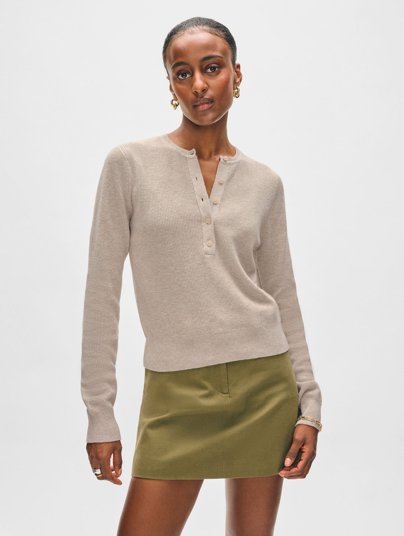 A woman with short hair and hoop earrings wears a White + Warren Fine Cotton Blend Henley sweater and an olive green mini skirt against a plain background, embodying versatile staple style with refined sophistication.