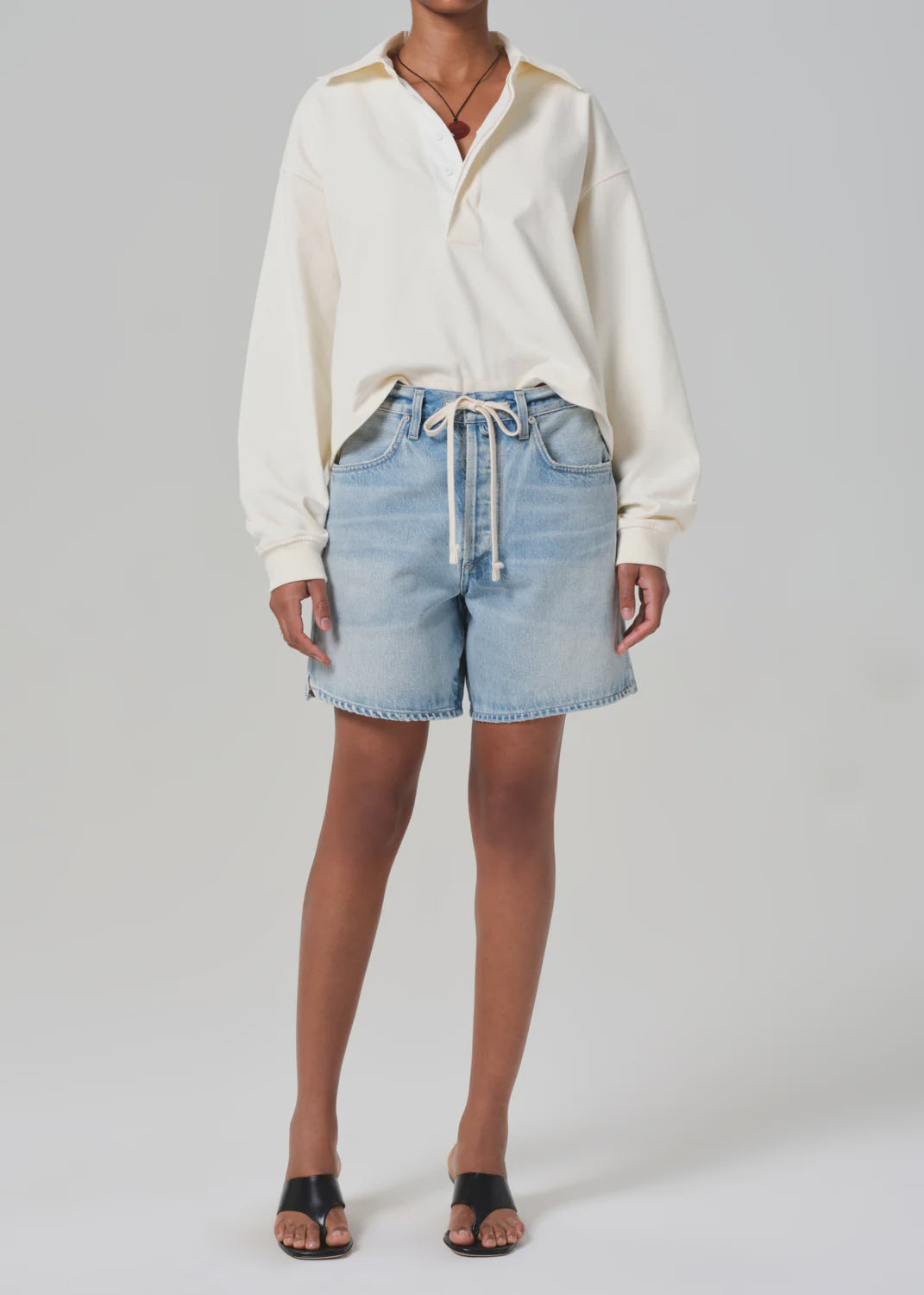 A person wearing a cream-colored long-sleeve shirt with Citizens Of Humanity Brynn Shorts in light blue, featuring a rope tie and crafted from regenerative cotton, paired with black open-toe sandals. All set on a plain gray background. Model size: 27.