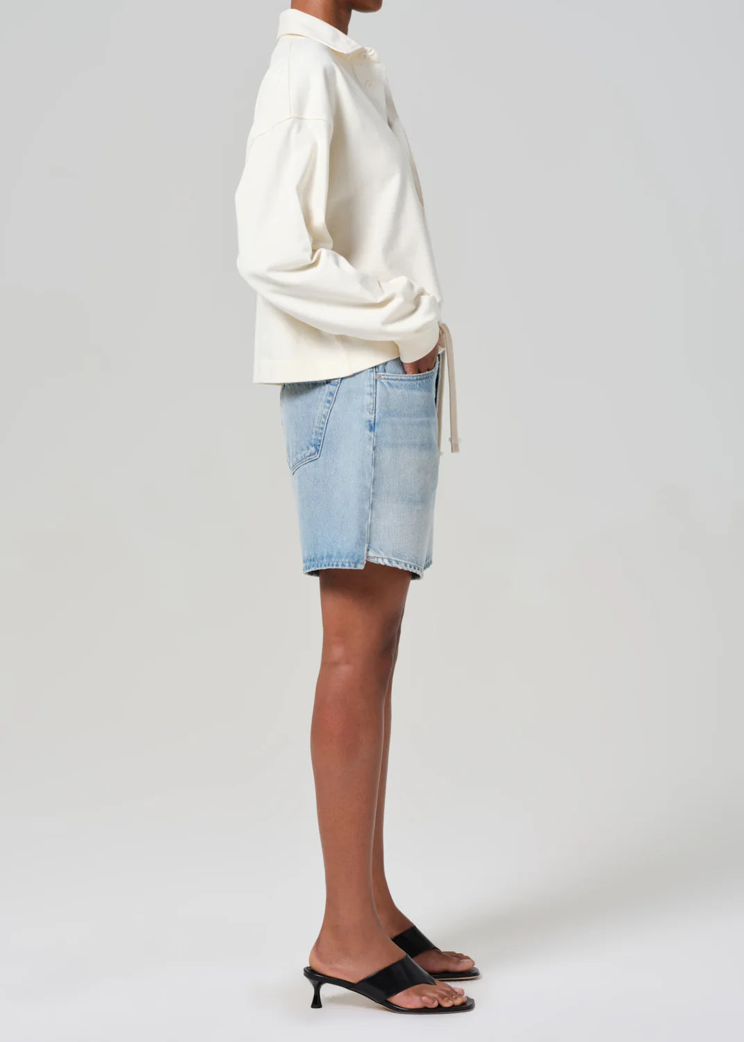 A person stands sideways against a plain background, wearing Brynn Shorts from Citizens Of Humanity/AGOLDE, a cream long-sleeve shirt, and black heeled sandals. The shorts are light blue denim made of breathable non-stretch fabric.