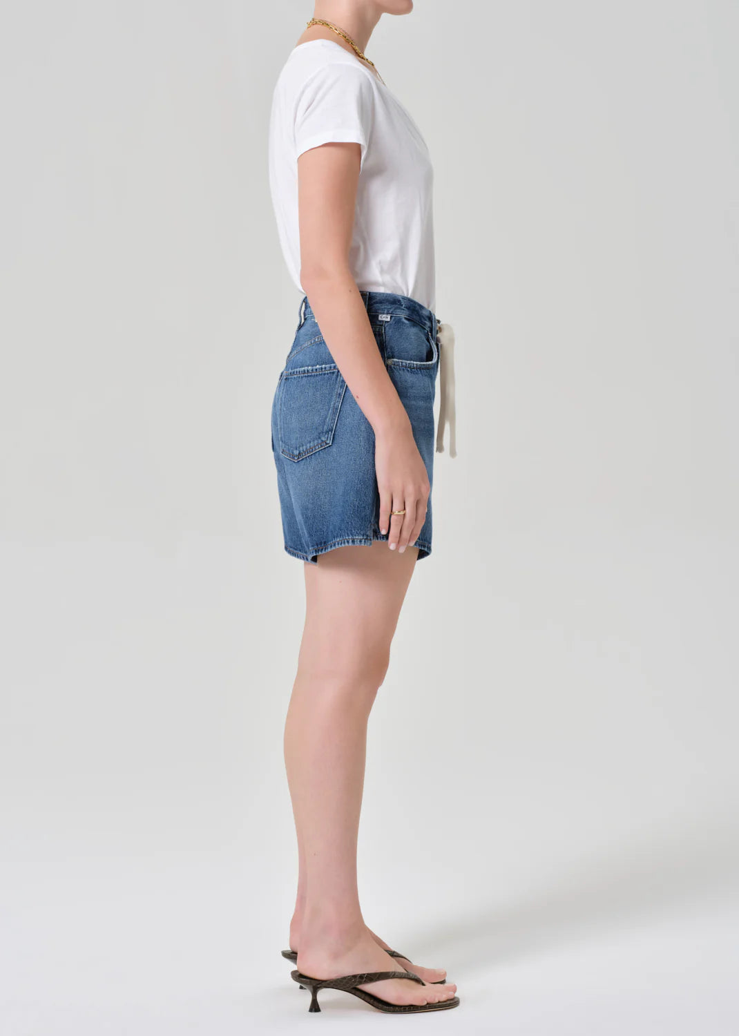 A woman stands sideways against a plain light background, wearing Citizens Of Humanity Brynn drawstring shorts in avant (lightwt vintage), a white t-shirt, and black flip-flops. Her hair is up and arms are relaxed by her side.