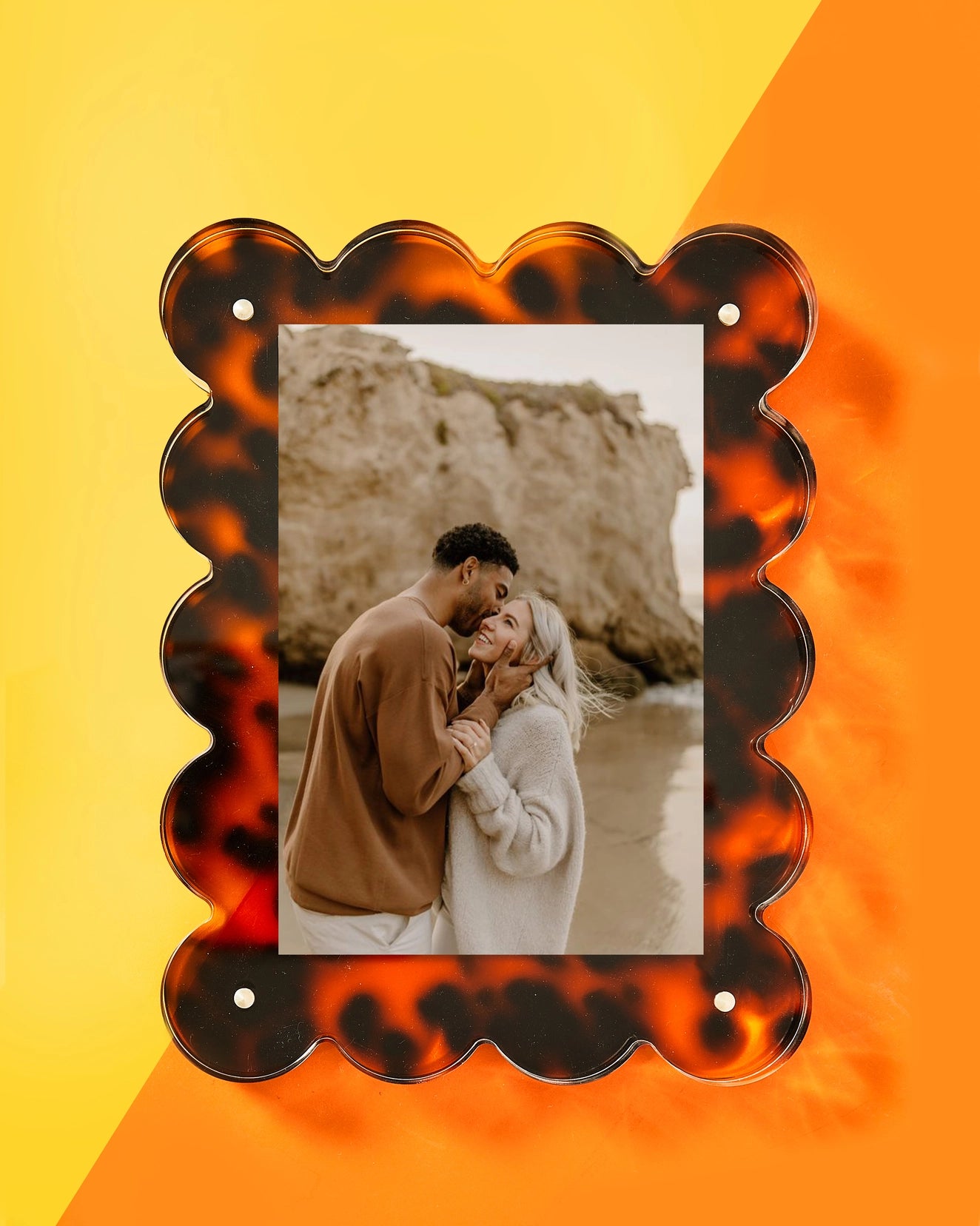 A couple embraces on a beach with rocky cliffs while smiling at each other. Their photo is displayed in Faire&#39;s decorative Acrylic Frame, featuring a scalloped tortoiseshell pattern, set against a bright yellow and orange background.