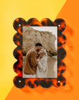 A couple embraces on a beach with rocky cliffs while smiling at each other. Their photo is displayed in Faire's decorative Acrylic Frame, featuring a scalloped tortoiseshell pattern, set against a bright yellow and orange background.