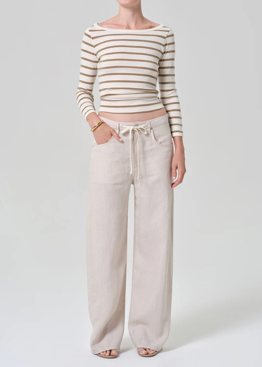 A person wears the Citizens Of Humanity/AGOLDE brynn drawstring trouser in linen—light beige with a relaxed fit and drawstring waist—paired with a beige and white striped long-sleeve crop top, standing against a plain light background.