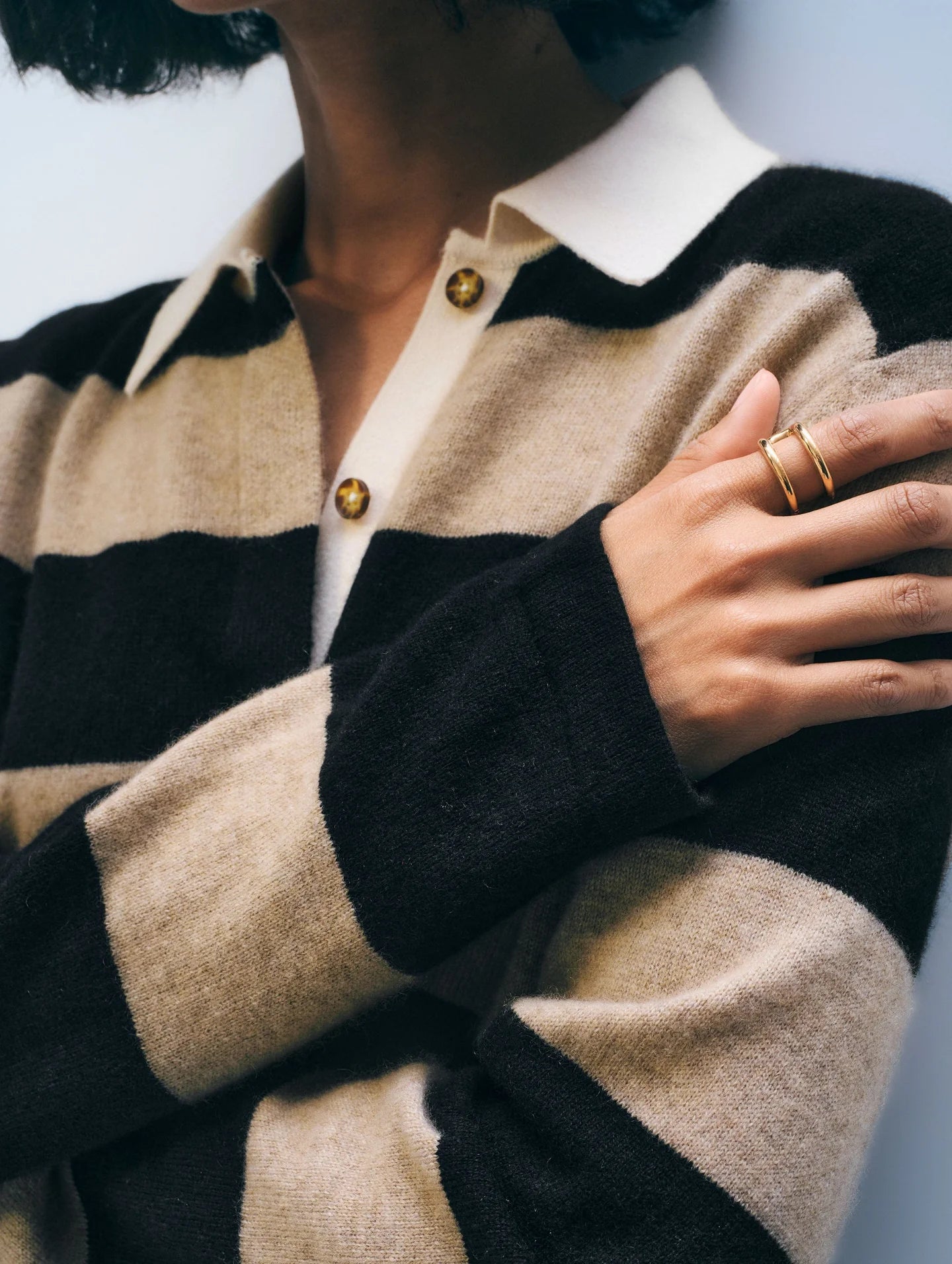 Wearing the White + Warren Cashmere Rugby Stripe Polo in beige and black, a person with chin-length dark hair crosses their arm to reveal gold rings. Gold buttons accent the sweater; their face is partially out of frame.