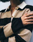 Wearing the White + Warren Cashmere Rugby Stripe Polo in beige and black, a person with chin-length dark hair crosses their arm to reveal gold rings. Gold buttons accent the sweater; their face is partially out of frame.