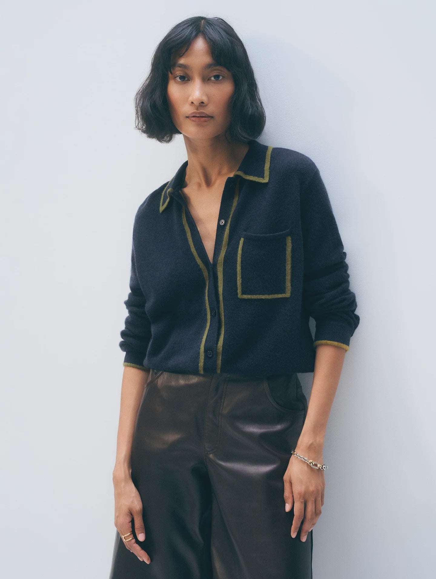 A woman with short, wavy black hair wears the White + Warren Cashmere Tipped Button Shirt in navy with green trim and front pocket, paired with high-waisted brown leather pants. She stands against a light background, looking calmly at the camera.