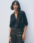 A woman with short, wavy black hair wears the White + Warren Cashmere Tipped Button Shirt in navy with green trim and front pocket, paired with high-waisted brown leather pants. She stands against a light background, looking calmly at the camera.
