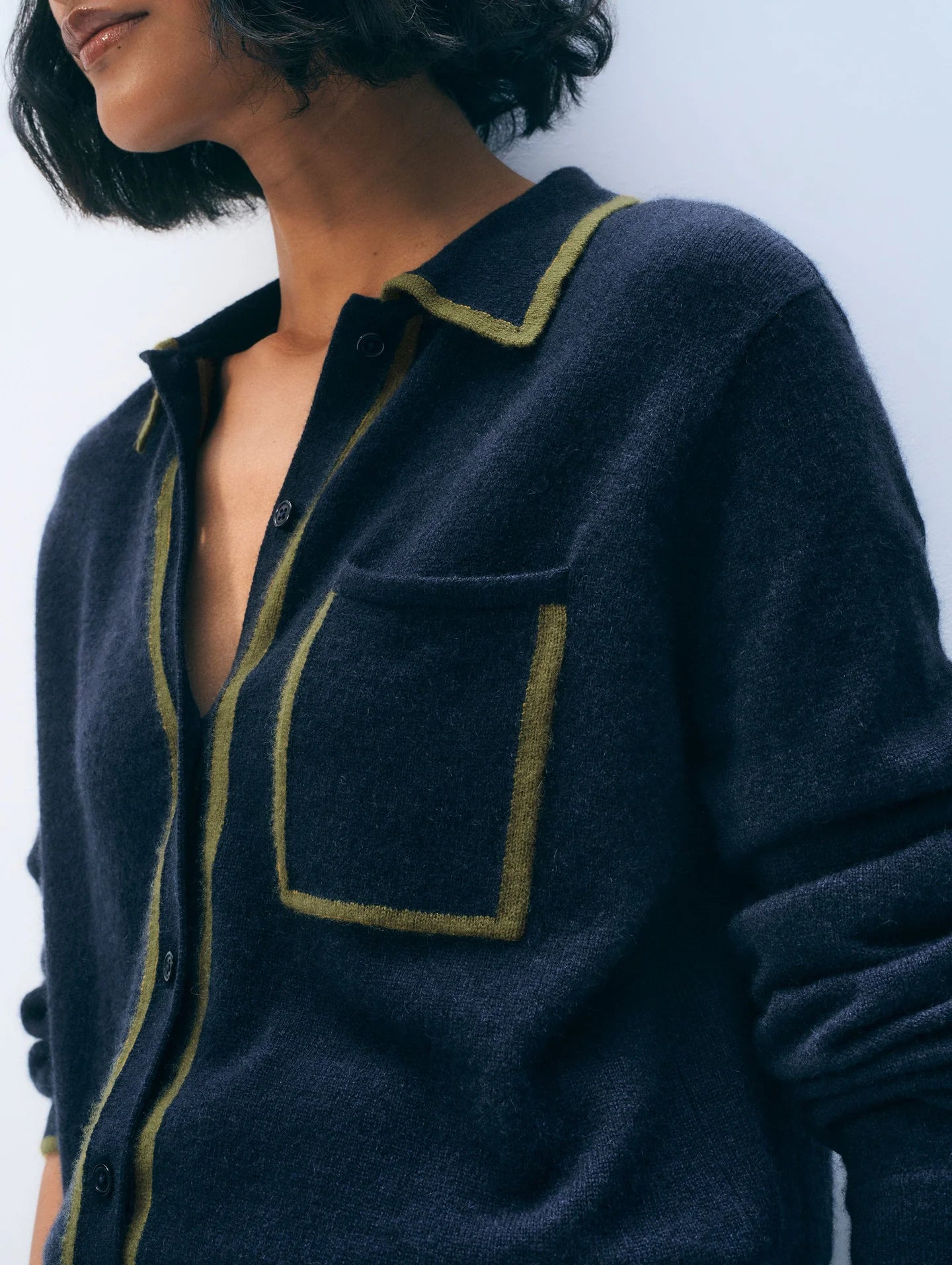 A person with a short, wavy bob wears the White + Warren Cashmere Tipped Button Shirt—a menswear-inspired dark blue cardigan with lime green trim at the collar, pocket, and front opening. The image is cropped at the shoulder and mouth.