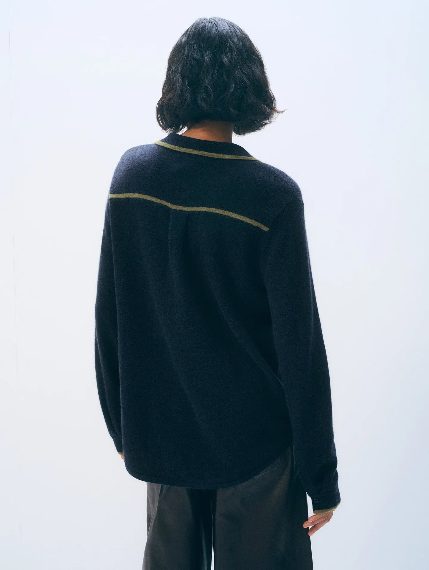 A person with short, wavy dark hair stands with their back to the camera, wearing the White + Warren Cashmere Tipped Button Shirt— a menswear-inspired, loose-fitting long-sleeve sweater with a horizontal stripe—paired with dark pants.