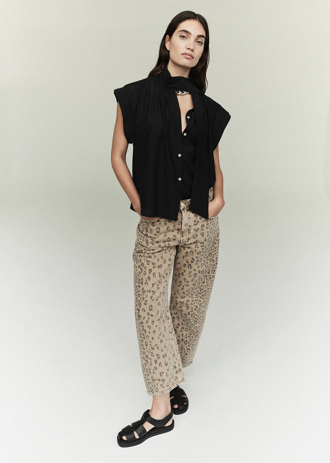 A woman with long dark hair and a neutral expression wears the Miro off white barrel leg wide-leg denim pants by Citizens Of Humanity/AGOLDE, paired with a sleeveless black top, scarf, and black sandals against a light background.