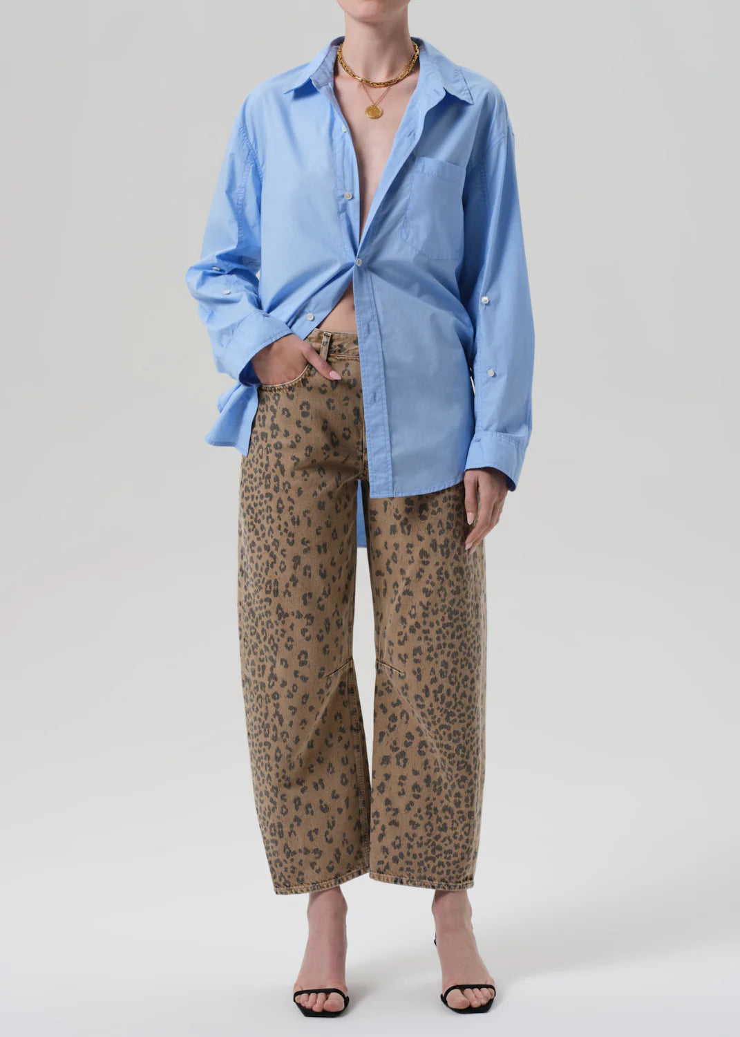 A person in the Miro shirt by Citizens Of Humanity/AGOLDE, loose light blue button-up, paired with off-white barrel leg jeans featuring black leopard print, black open-toe heels, and a gold necklace against a plain light backdrop.