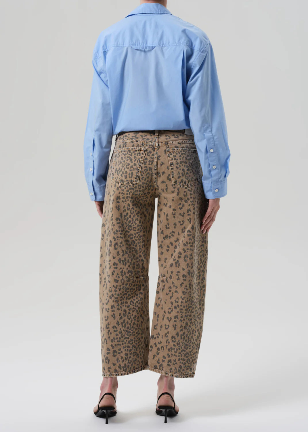 A person faces away, wearing the Miro off white barrel leg leopard print denim pants by Citizens Of Humanity/AGOLDE, paired with a light blue button-up shirt and black heeled sandals against a plain background.