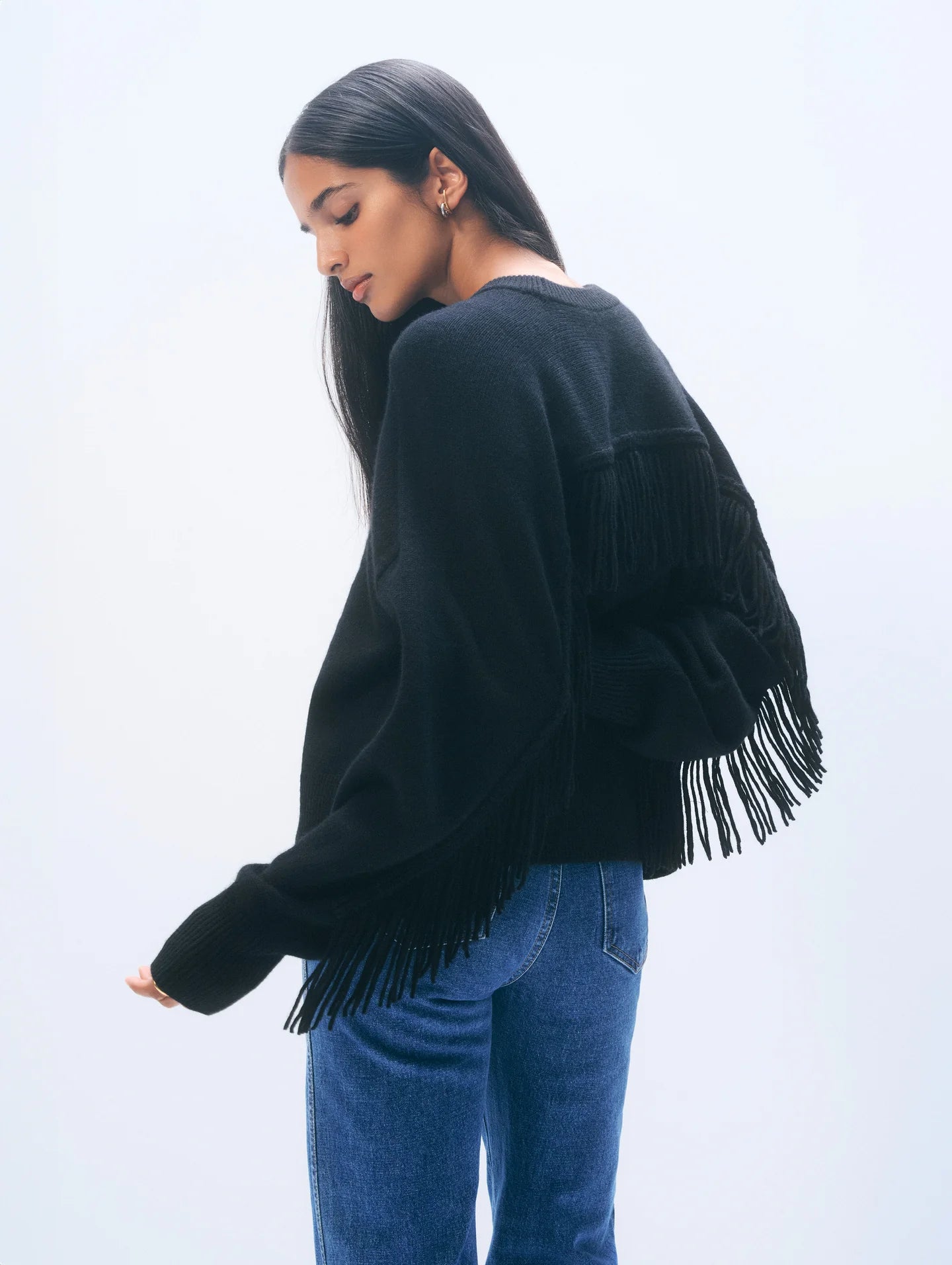 A woman with long dark hair wears the White + Warren Cashmere Luxe Fringe Crew, an oversized black sweater with fringe on the back and sleeves, styled with blue jeans. She stands sideways against a light, plain background, looking down.