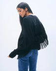 A woman with long dark hair wears the White + Warren Cashmere Luxe Fringe Crew, an oversized black sweater with fringe on the back and sleeves, styled with blue jeans. She stands sideways against a light, plain background, looking down.