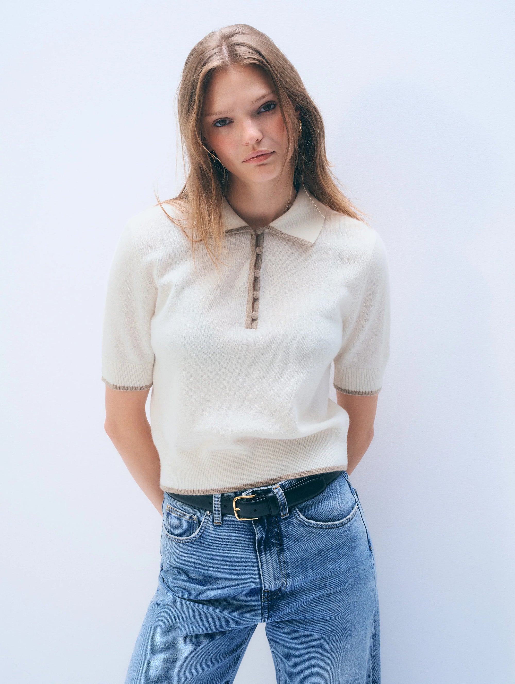 A woman with straight, light brown hair wears the White + Warren Cashmere Button Down Polo in cream, paired with blue jeans and a black belt. She stands against a plain white background, looking seriously at the camera.