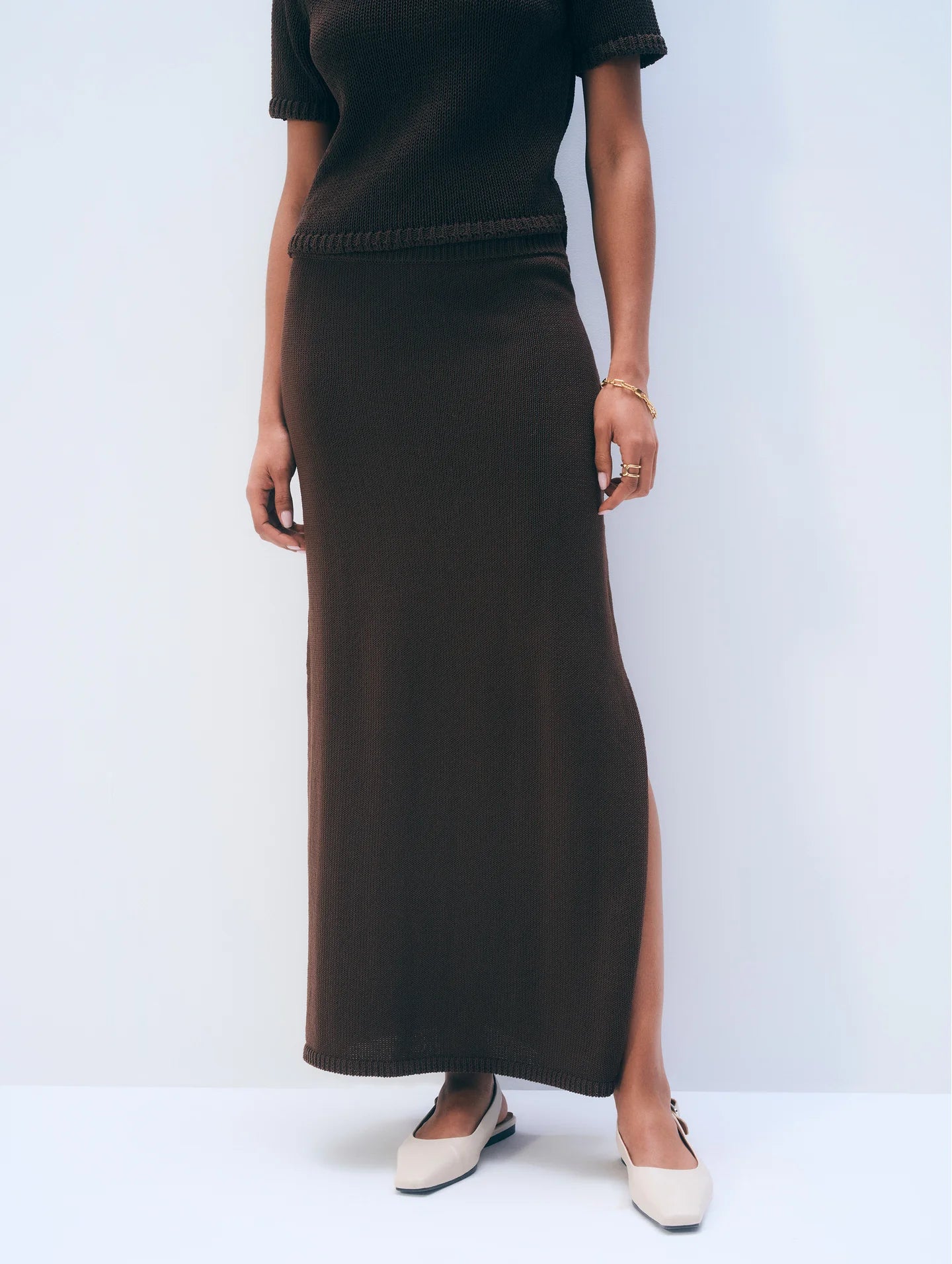 A person stands against a plain background wearing the Luxe Cotton Skirt by White + Warren in dark brown with a matching short-sleeve top featuring a side slit, styled with white pointed-toe flats and a delicate bracelet.