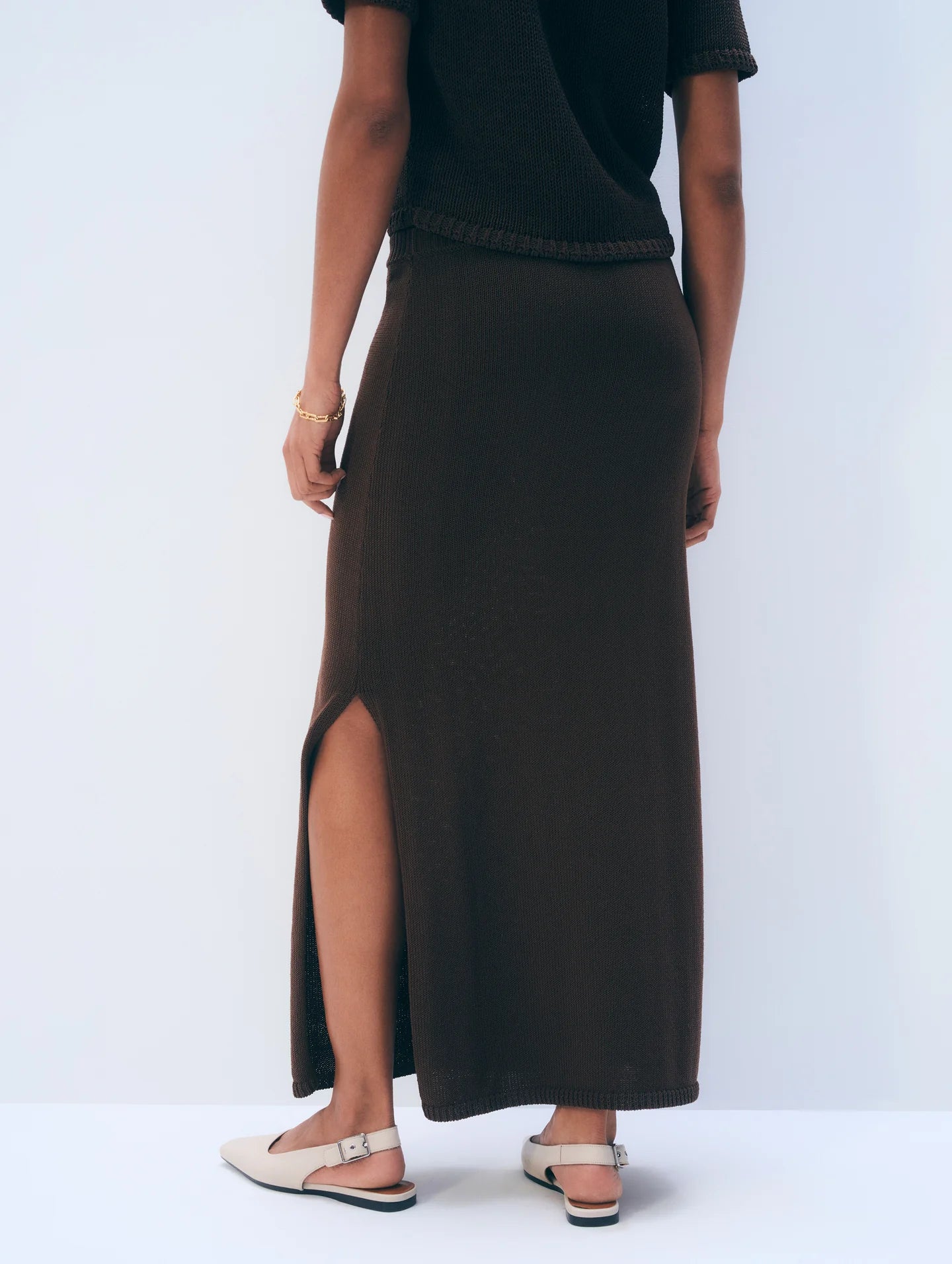 A person stands against a light background, shown from the back, wearing the White + Warren Luxe Cotton Skirt in dark brown with a side slit, paired with a matching top, gold bracelet, and beige slingback flats.