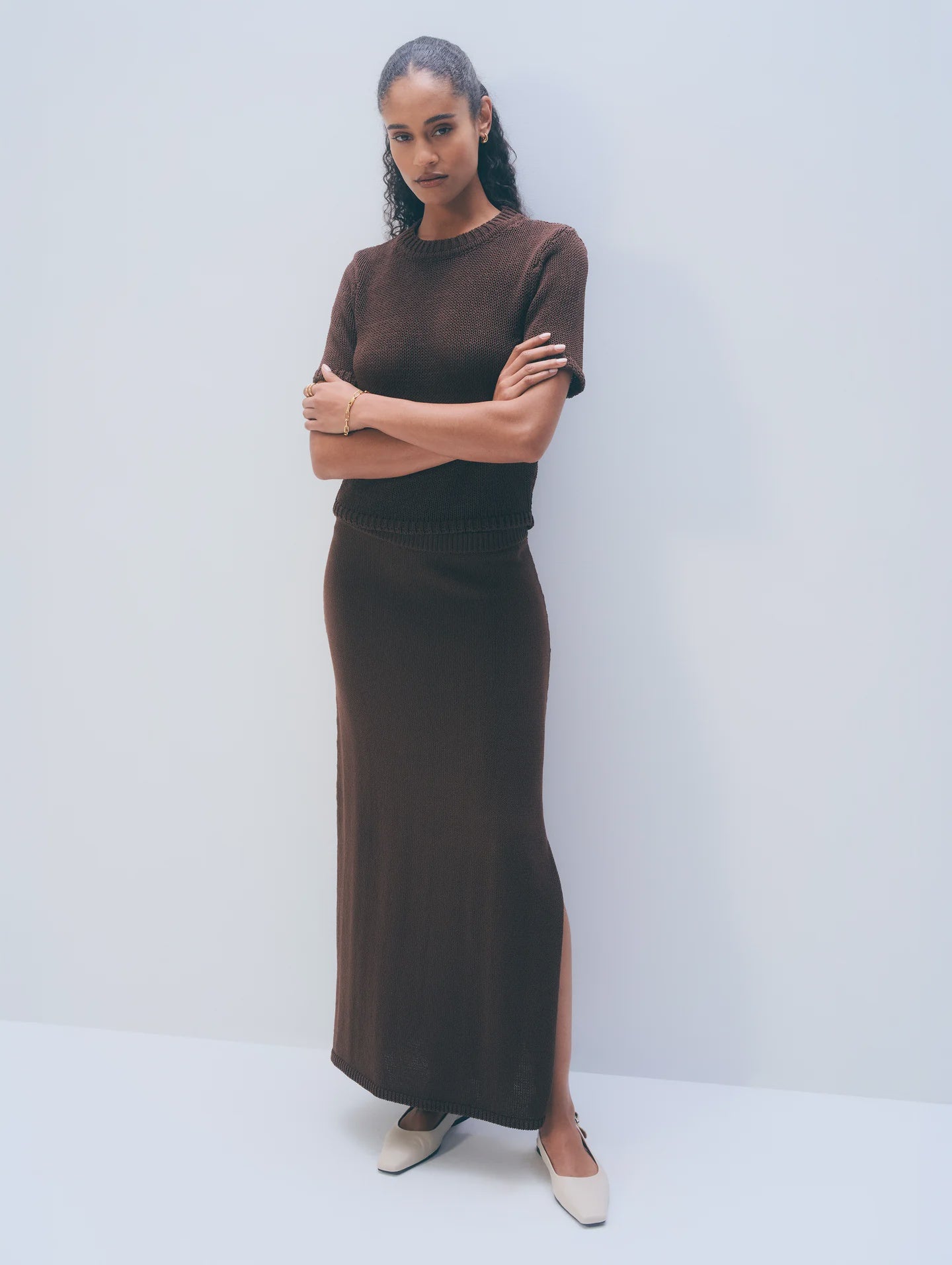 A woman with curly hair stands against a plain background, arms crossed, wearing White + Warren's Luxe Cotton Skirt in dark brown with a side slit and a matching Italian knit top. She completes the look with light pointed shoes.