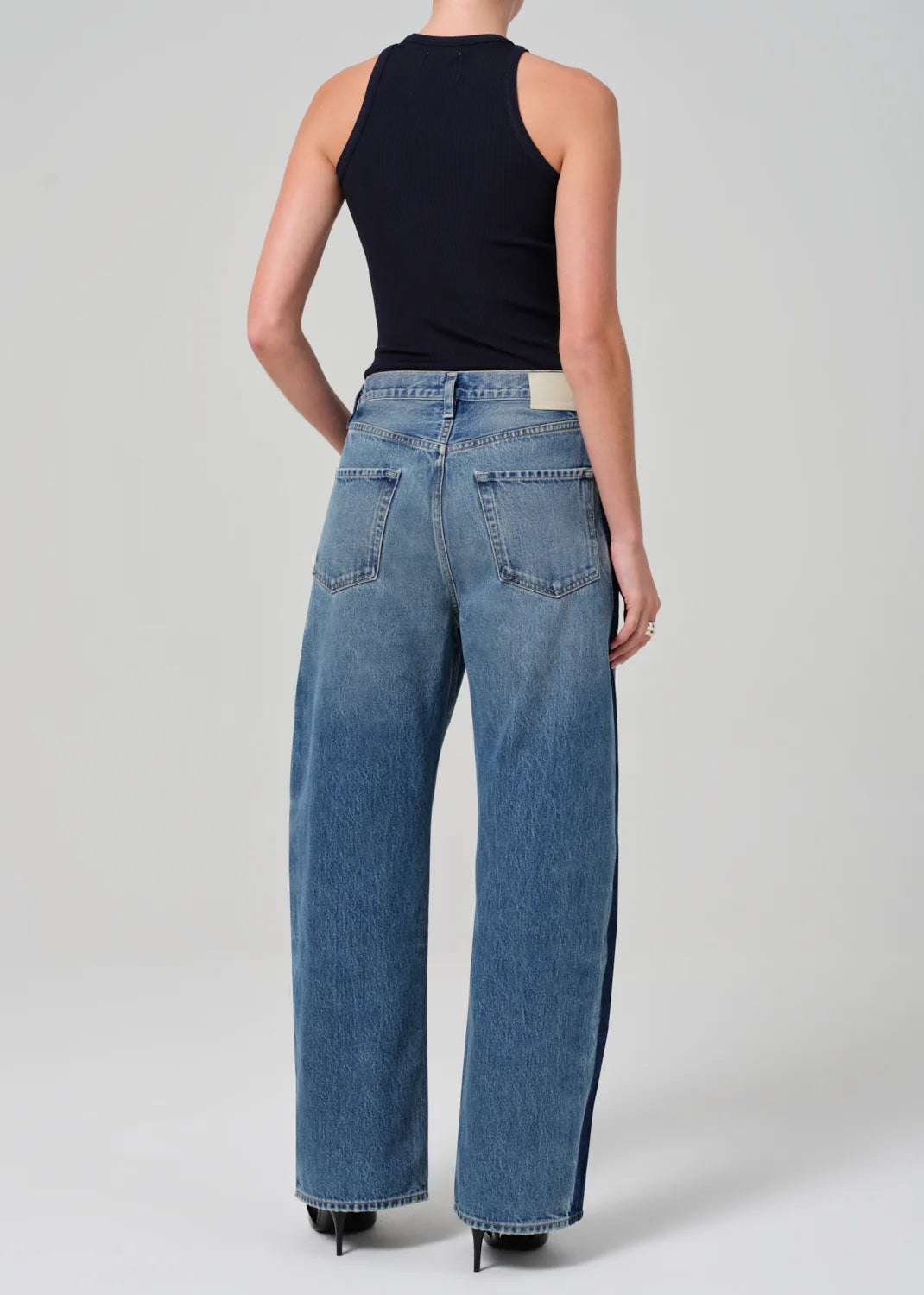 A person wearing a black sleeveless top and the Ayla Baggy Tuxedo Stripe jeans from Citizens Of Humanity/AGOLDE stands facing away from the camera. These tailoring-inspired, high-waisted jeans, crafted from regenerative cotton, feature a loose fit and two back pockets against a plain light gray background.