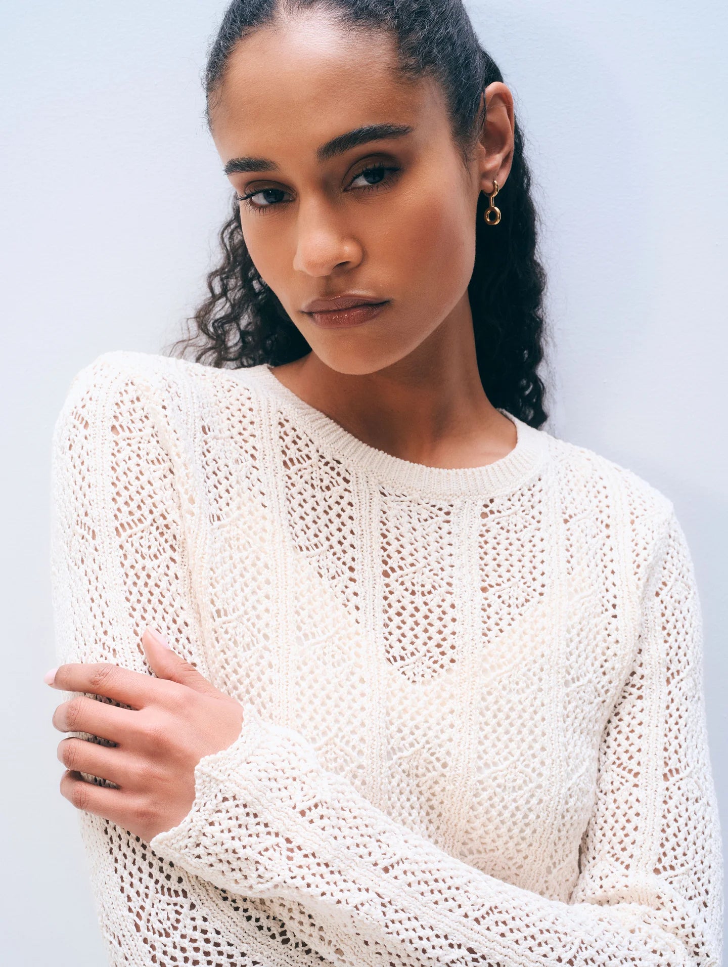 A woman with long curly hair wears the White + Warren Cotton Pointelle Top and small hoop earrings, standing against a plain light background with a confident, neutral expression.
