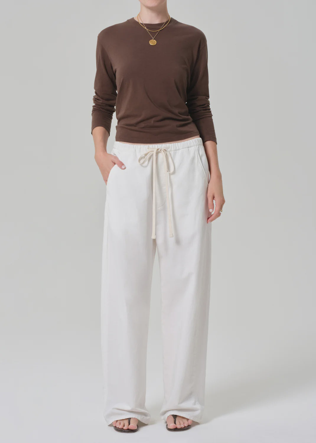 A person stands against a plain background wearing a brown long-sleeve shirt, Citizens Of Humanity/AGOLDE aurora pull on lightweight summer pants in an off-white cotton flax blend, black sandals, and layered gold necklaces.