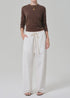 A person stands against a plain background wearing a brown long-sleeve shirt, Citizens Of Humanity/AGOLDE aurora pull on lightweight summer pants in an off-white cotton flax blend, black sandals, and layered gold necklaces.