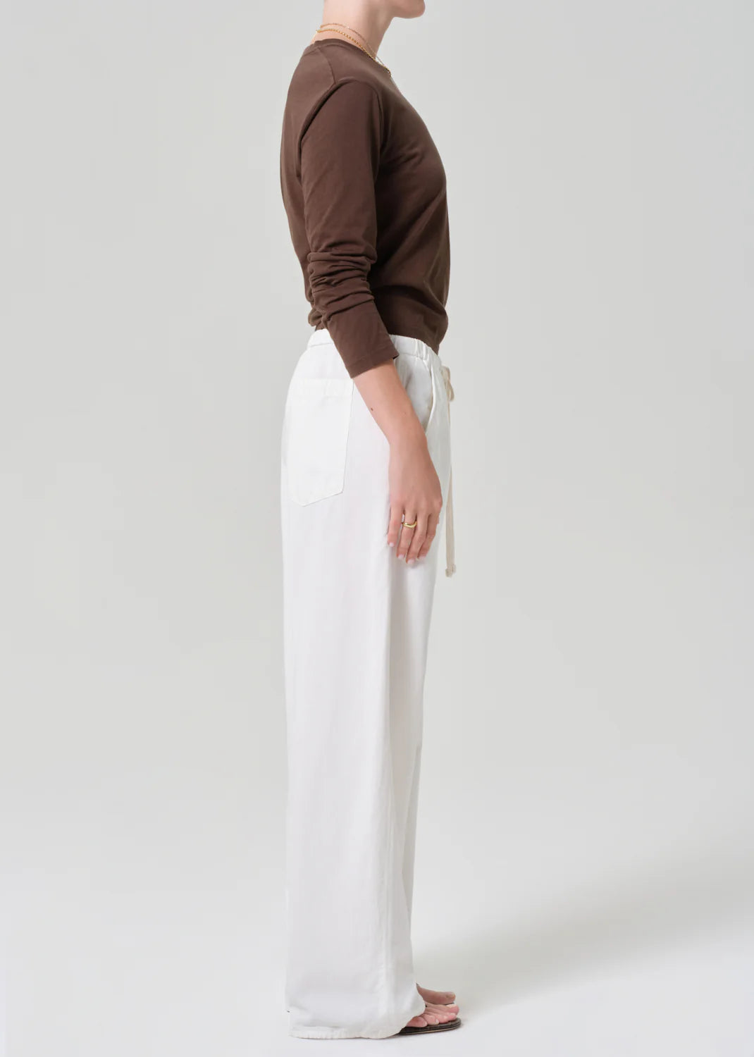 A person stands sideways against a plain background wearing a long-sleeve brown shirt and the Citizens Of Humanity/AGOLDE aurora pull on pant—a loose-fitting, soft cotton flax blend women’s pant with front pockets. Their left arm is relaxed, feet bare.