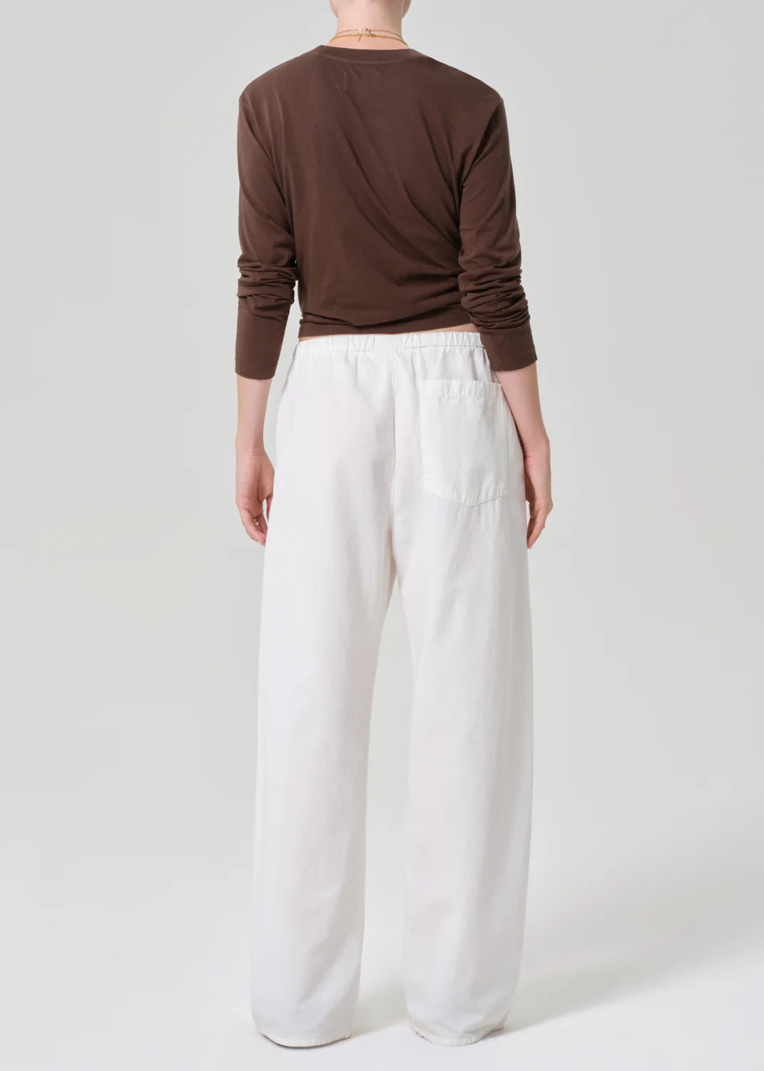 A person stands facing away, wearing a long-sleeved brown top and the Citizens Of Humanity/AGOLDE aurora pull on pant—loose-fitting with an elastic waistband in a cotton flax blend—against a plain light gray background.