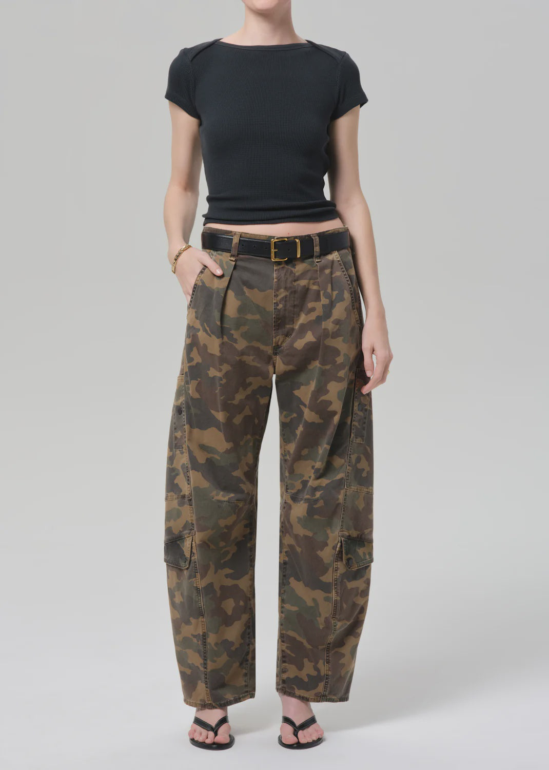 A person wears a fitted black crop top with Citizens Of Humanity/AGOLDE flight pant in camo/khaki, styled with a black belt, black sandals, and a gold bracelet, standing against a plain light gray background.