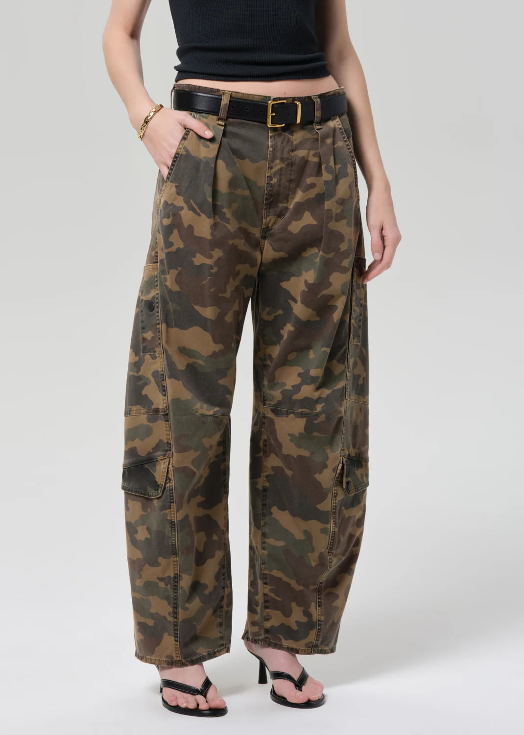 A person stands with one hand in a pocket against a plain background, wearing Citizens Of Humanity/AGOLDE flight pant in camo/Khaki, a black belt with gold buckle, black crop top, gold bracelet, and black heeled sandals.